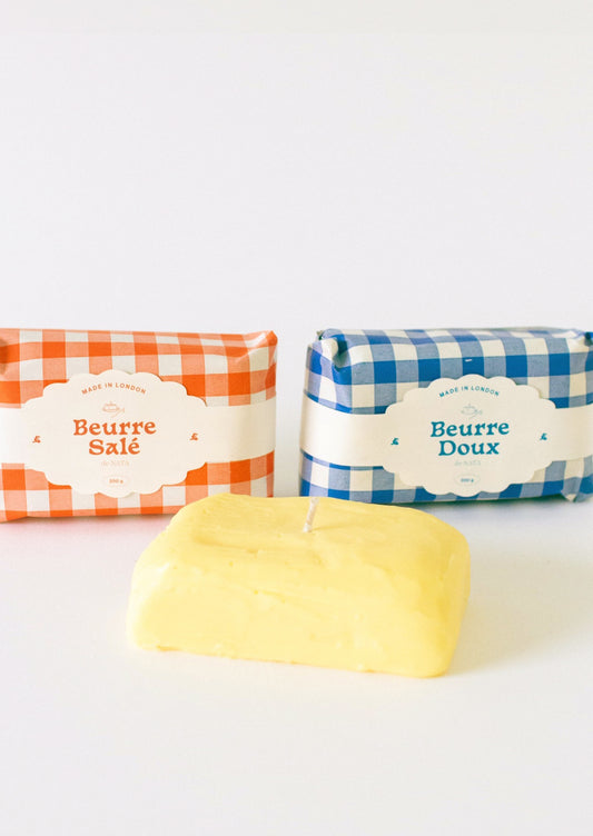 A candle in the shape of a block of butter, gingham packaging in red or blue.